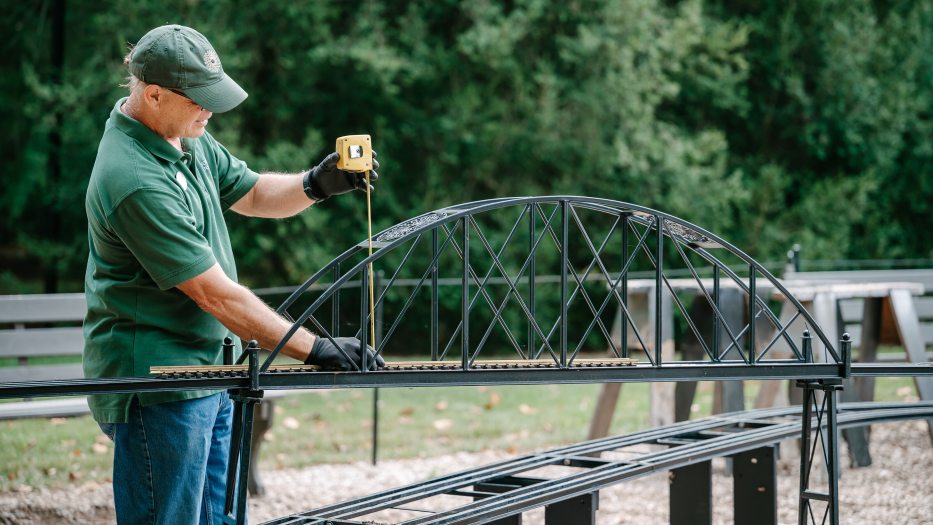 A person in a green Longwood Gardens shirt and cap extends a tape measure from a miniature outdoor railway track to the overhead arch of a bridge.