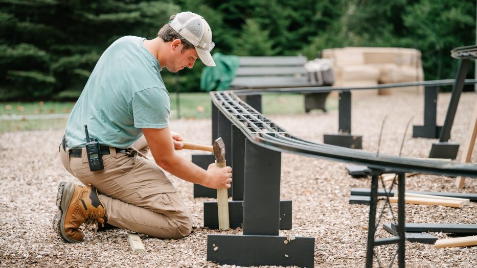 A kneeling worker hammers a stake into mulched ground to help support an outdoor miniature railway.