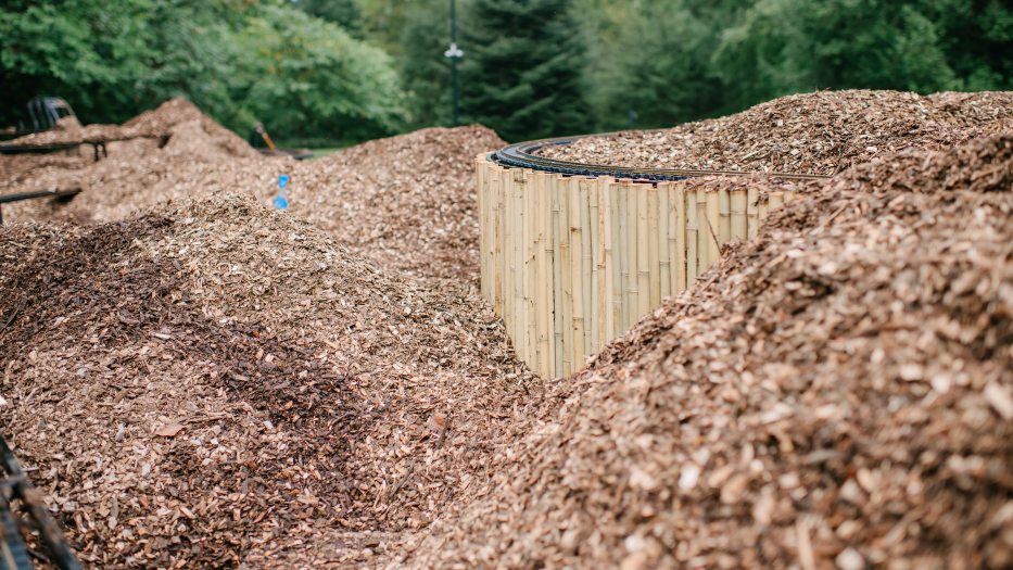 A curving miniature train track next to a curved bamboo fence peek out from undulating piles of light brown mulch.