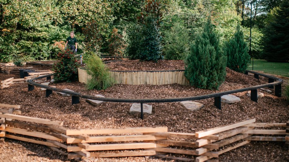 A curving miniature train track runs through a landscape of trees, stepping stones, mulch, and fencing.