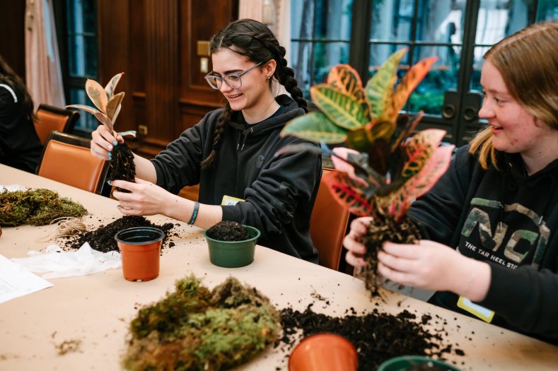 two teens holding small plants and breaking up the dirt around the roots