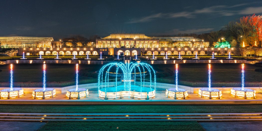 Fountains of light mimic water and flame in the foreground, with an expansive and warmly lit conservatory complex in the background, against a dark blue sky.
