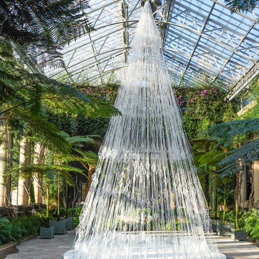 A tall tree shaped from white strands of thread strung with white ornament shapes, against a green backdrop and high glass ceiling.