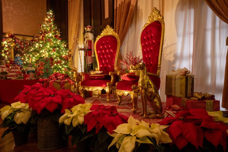 Two red thrones topped with pillows and bejeweled crowns are flanked by gold dogs, red and white poinsettias, with a table of decorated cakes and an ornate Christmas tree and garland to the left.