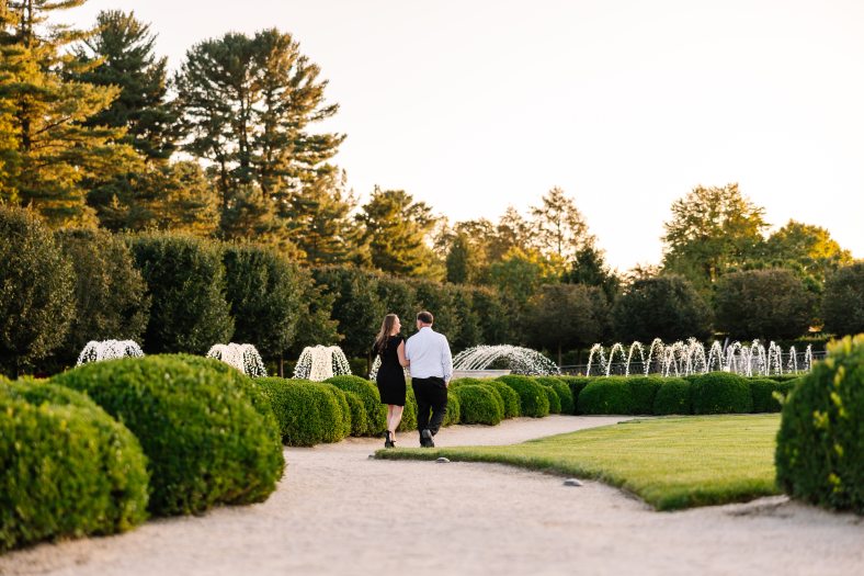  A couple walks along a gravel path in a garden with green bushes and fountains under a clear sky.
