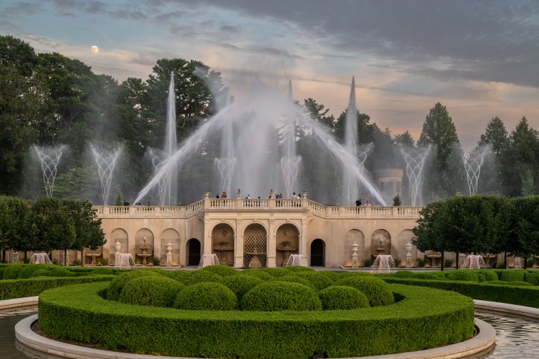 Fountain jets soar high above a fountain with a circular section of green boxwood hedges in front of it.