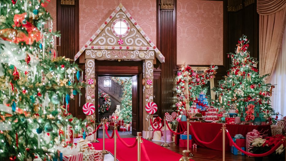 A festive holiday display featuring a large gingerbread house entrance flanked by multiple decorated Christmas trees and oversized candy canes, set in an ornately wallpapered room.