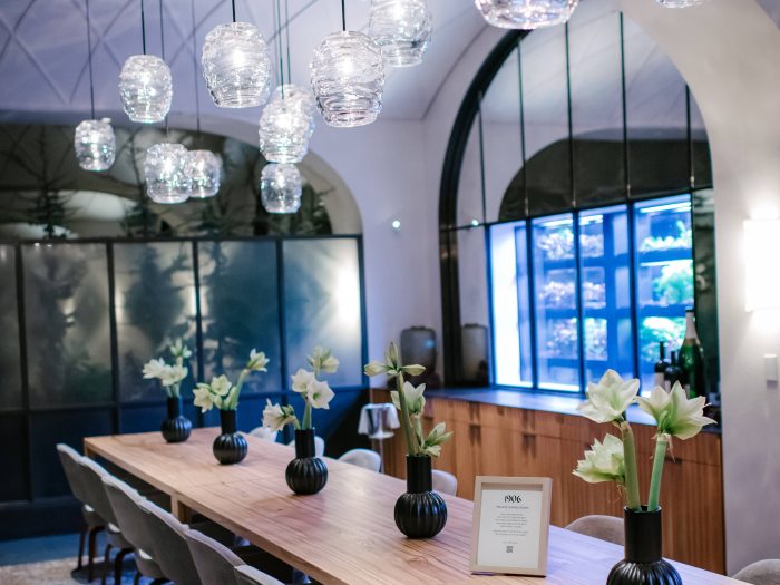 Long wooden dining table adorned with flower vases, with plush gray chairs and overhanging globe lights.