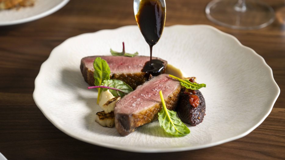 Two slices of duck breast on a white plate, with a spoon dripping a dark brown sauce onto the meat.