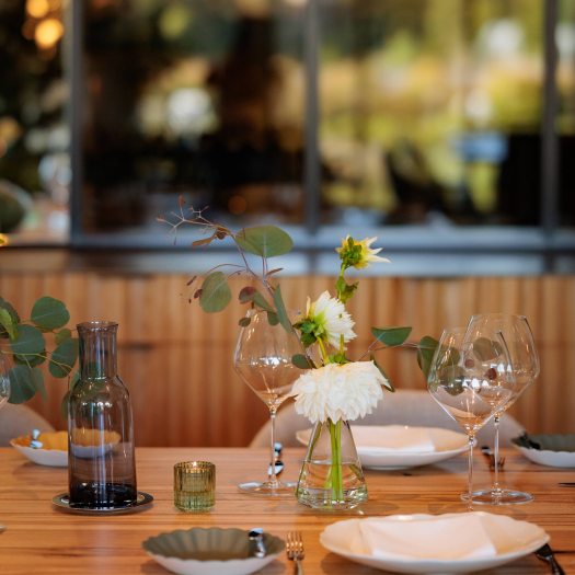 Table setting with white plates and flowers, wine glasses, votive candle, and glass decanter; with large window in background.