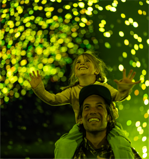 A joyfully smiling child rides on the shoulders of a smiling adult as they gaze upward at yellow-green sparks of light.