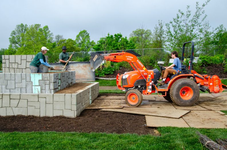 A person operating an orange tractor type machine to build a sculpture in a garden.
