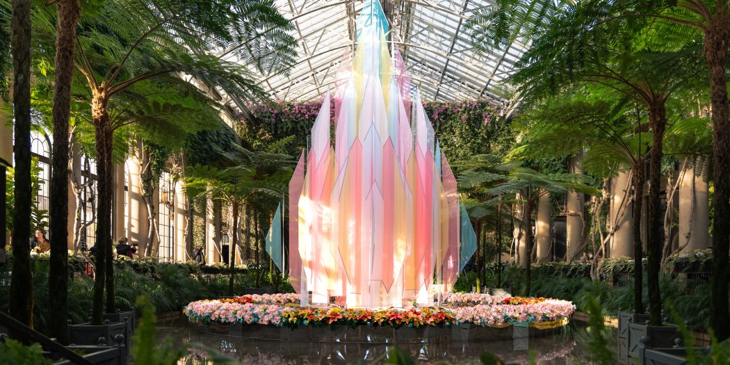 Geometric cloth panels in sheer pastel colors combine to form a diamond-shaped Christmas tree amid potted Christmas plants in a lush indoor conservatory setting.