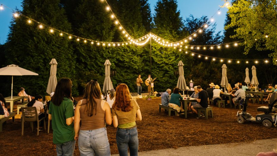 Guests stand or sit at outdoor tables, with strings of party lights leading to a small stage hosting live performers in the background.