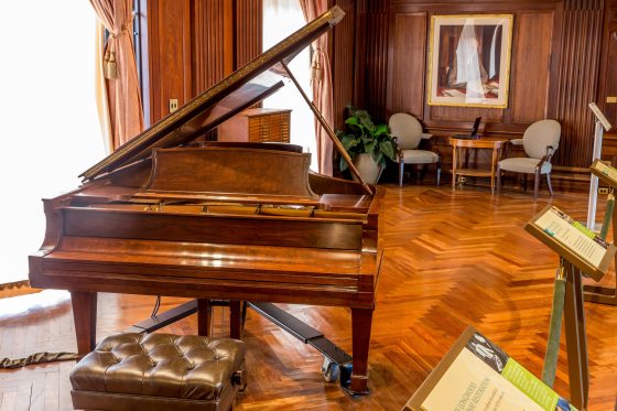 A large wooden piano sits in a room with a wooden floor and historic furniture from the early 1900s
