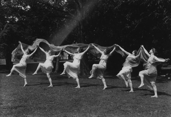 A line of women dressed in white strike a dance post with one foot in the air and holding hands down the line