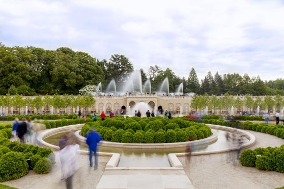 Visitors stroll through a fountain garden lush with green boxwoods and fountain jets shooting in the air