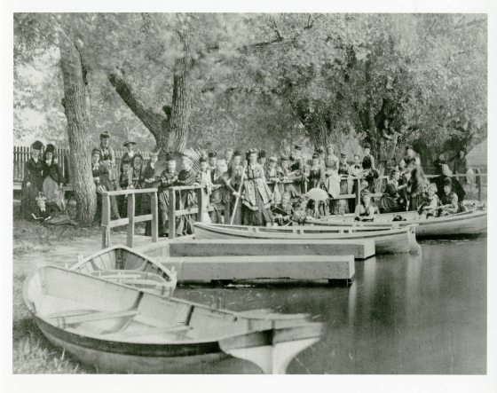 Historic photo of a large group of people gathered at the side of a small lake awaiting to go on rowboats