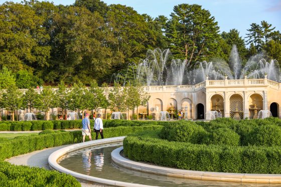 two people walk near a circular basin of water in the center of a large, ornate fountain garden