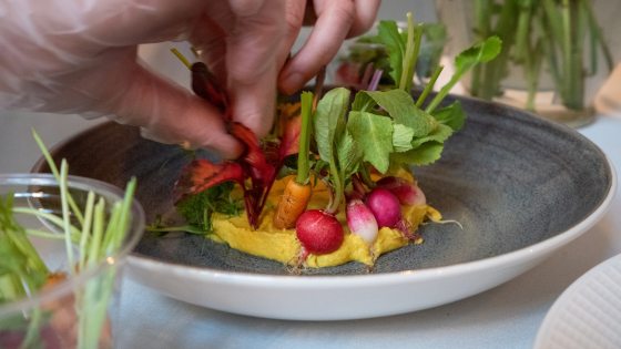 person placing small vegetables in a dish of yellow hummus