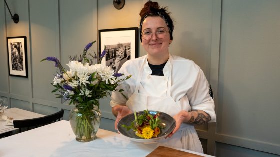 chef holding a plate of food from behind a table and a vase of flowers next to them