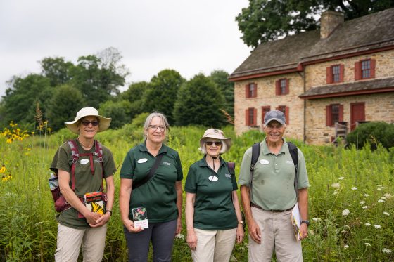 4 people in green shirts with backpacks standing in a meadow next to an old farmhouse