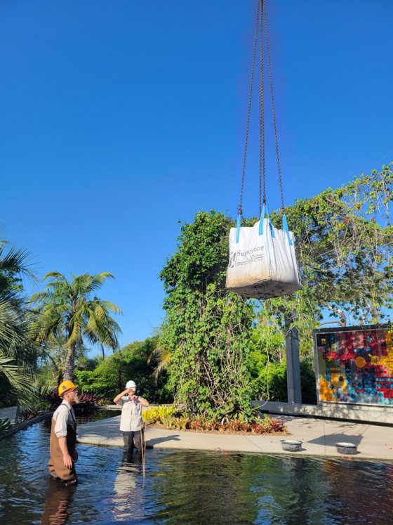 two people in hard hats standing in a pool of water in waders with a bag being lifted into the pool by a crane