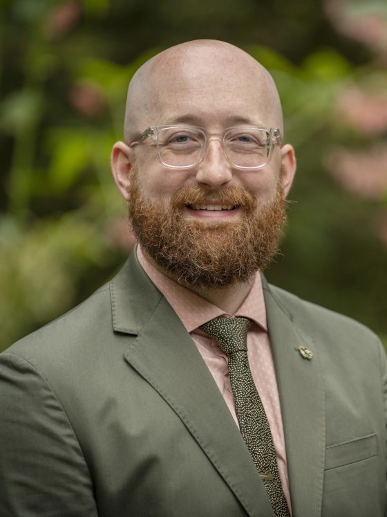 person in an olive green suit and clear glasses looking at the camera
