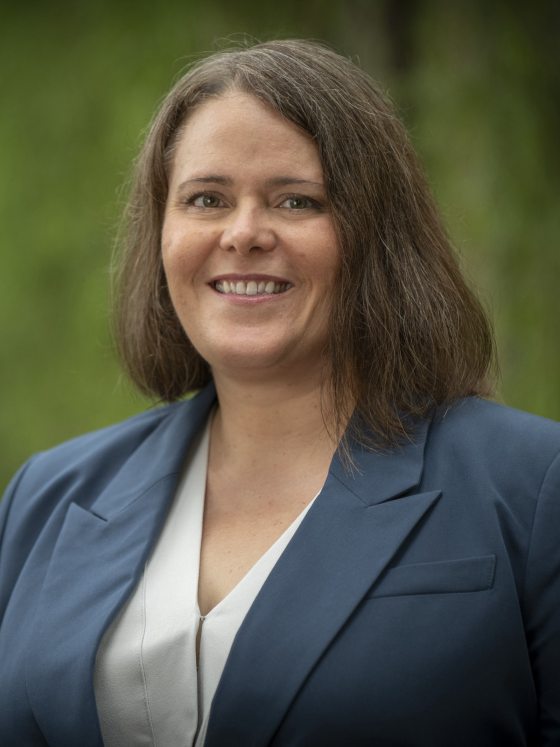 person with slate blue jacket over a white V-neck top smiles at the camera