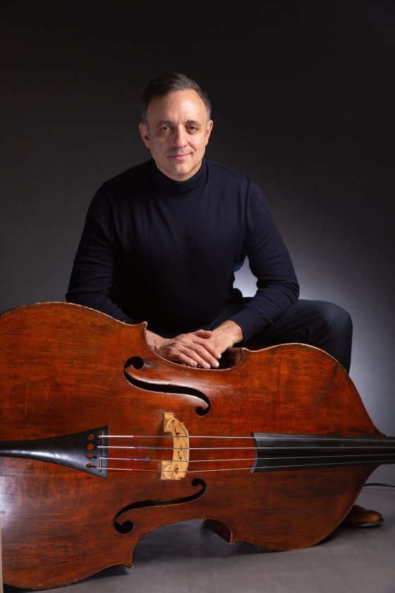 portrait of a musician dressed in black, kneeling behind a bass that is turned on its side