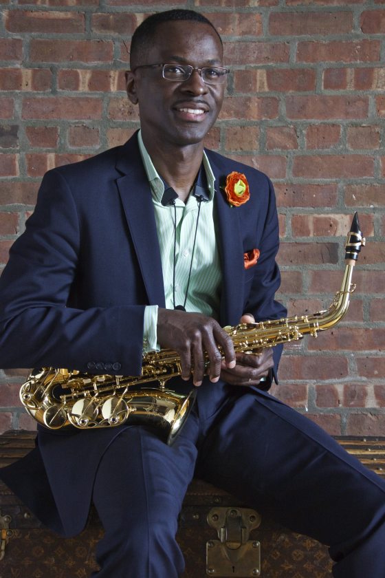 portrait of a seated musician—wearing a dark blue suit, green striped shirt, and matching orange boutonniere and pocket handkerchief—holding a saxophone