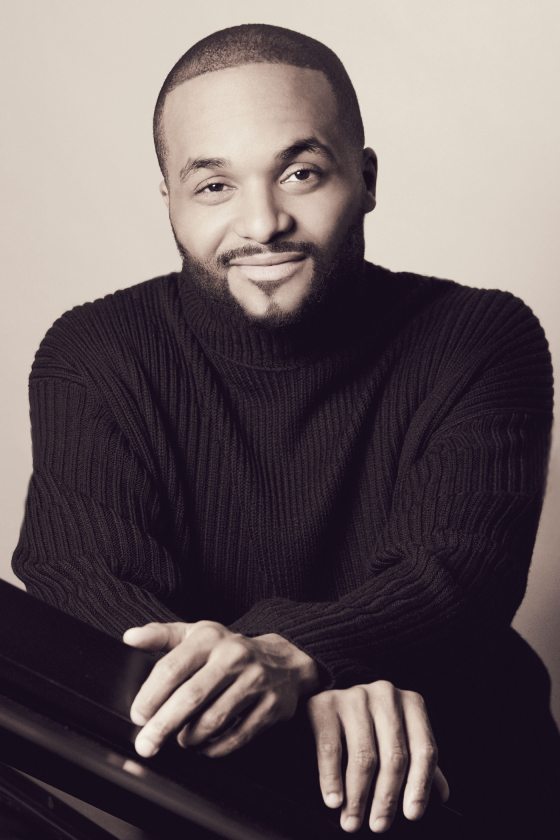 a musician dressed in black poses behind a piano
