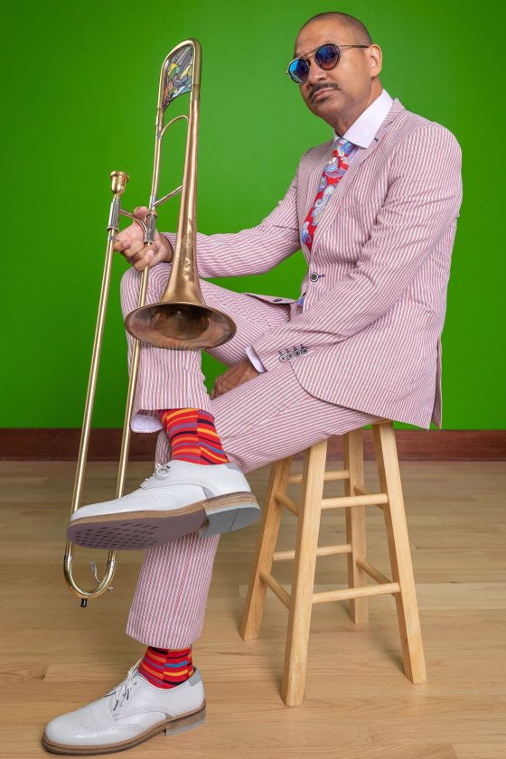 a seated musician—wearing a red and white striped suit; a red, white and blue paisley tie; brightly patterned red socks; and sunglasses—holds an ornate trombone