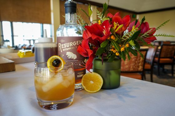 a citrus drink, bottle of whiskey, and a vase of flowers on a white table