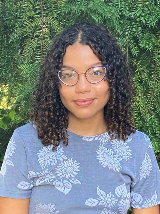 portrait of smiling person wearing glasses, with curly shoulder-length brown-black hair and blue floral shirt