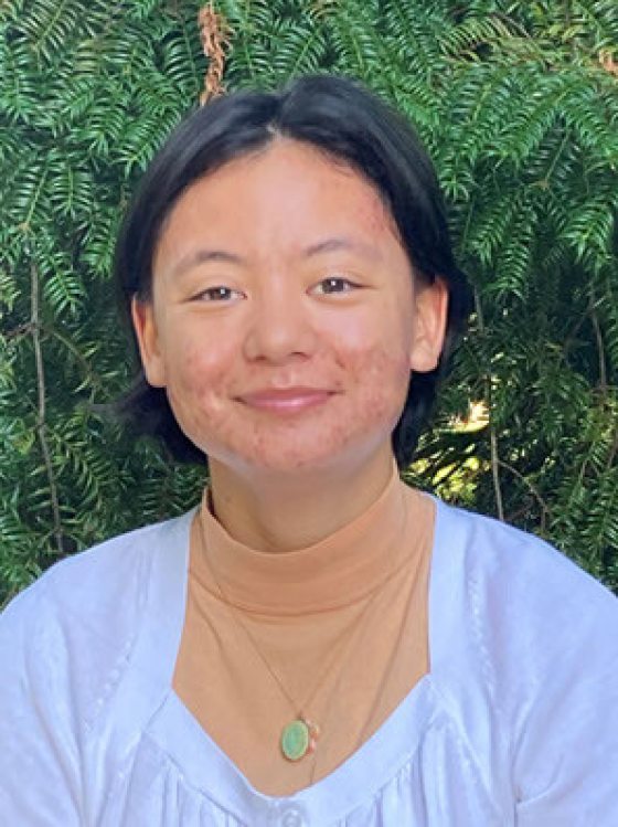 portrait of smiling person with short black hair, wearing a white top over a tan mock turtleneck