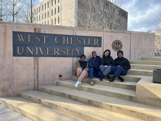 Four people outdoors sitting on stone steps with a sign behind them that read West Chester University