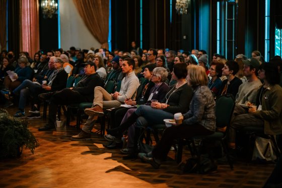 a dim room full of people seated in rows listing to a speaker