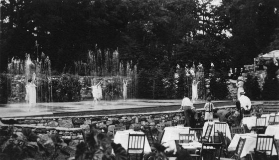 black and white image of dancers on a stage among fountains, while a few people casually observe from an audience filled with small dining tables