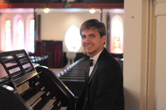 musician seated at church organ with pews and stained glass windows in background
