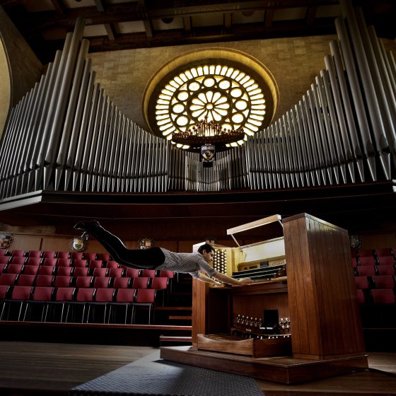 an organist appears to fly away while playing an organ console, against a backdrop of organ pipes and a large round stained glass window