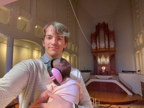 a person in a white sweather looking at the camera while holding a baby who is in pink earphones