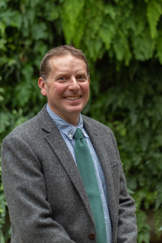 Colin Skelly poses next to Longwood's Green Wall with a grey suit and green tie.