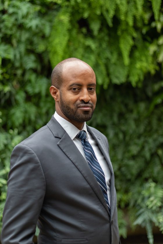 Muluken Kebede poses in a grey suit next to Longwood's Green Wall.