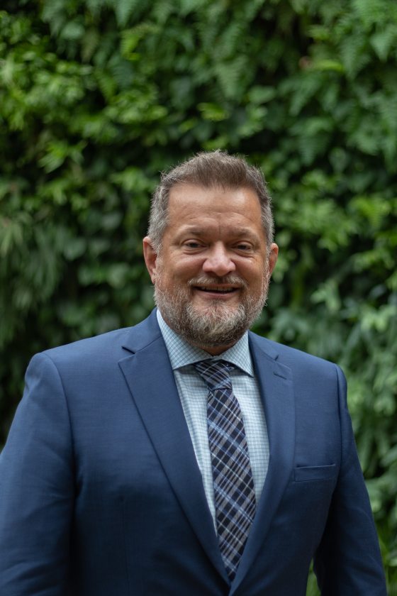 A man with a beard wearing a navy suit poses in front of Longwood's Green Wall.
