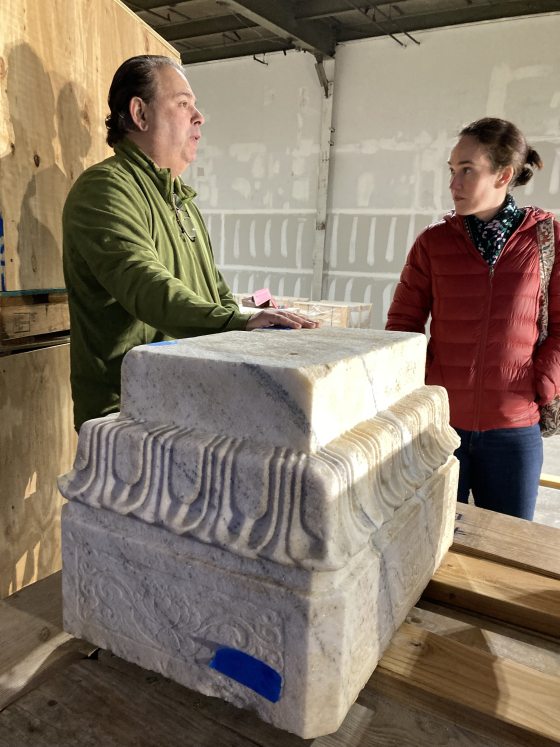 two people standing and talking behind a large stone sculpture base