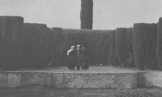 Three people stand on an outdoor stone stage in a garden in an old black and white photo from the early 20th century
