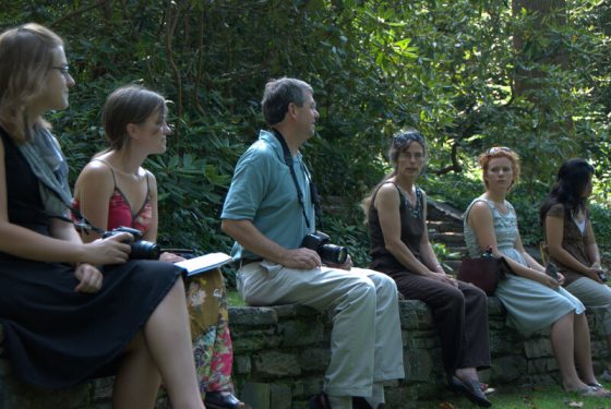 Six people sitting outdoors on a stone wall listening to one of them speak.