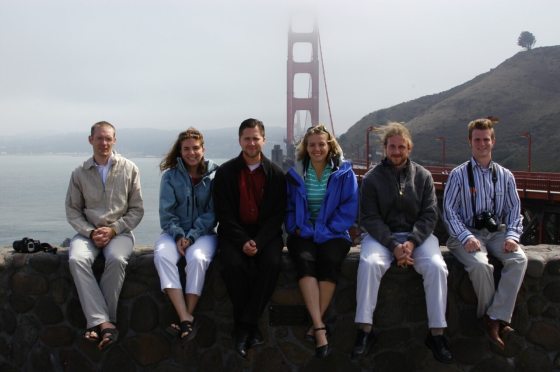 A group of six people sitting on a stone wall overlooking a body of water with a bridge in the background.