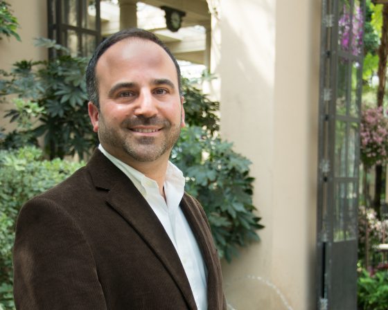 A person in a brown jacket and white shirt turned at an angle smiling at the camera.